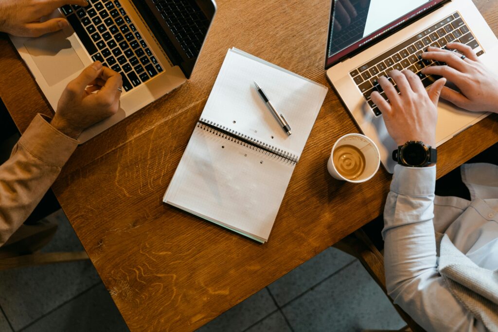 Overhead shot of coworkers using laptops with notes and coffee at a wooden desk.